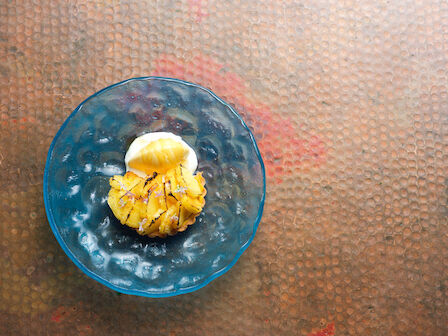 A blue plate with a dessert featuring a scoop of ice cream and grilled fruit, possibly pineapple, on a textured brown surface.