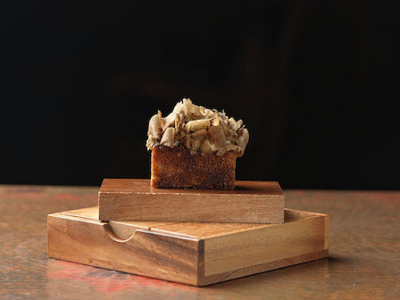 A small rectangular cake with frosting sits on a wooden box against a dark background, creating an artistic, minimalist presentation.