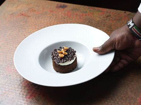 A hand presenting a dessert on a white plate, topped with chocolate and nuts, placed on a textured surface.