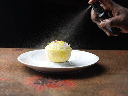 A person sprays a mist over a decorative dish with intricate garnishes on a white plate, set against a dark background.