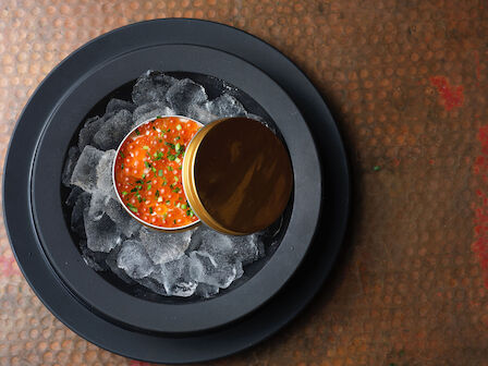 The image shows a tin of red caviar garnished with herbs, placed on a bed of ice in a black dish, against a textured surface.